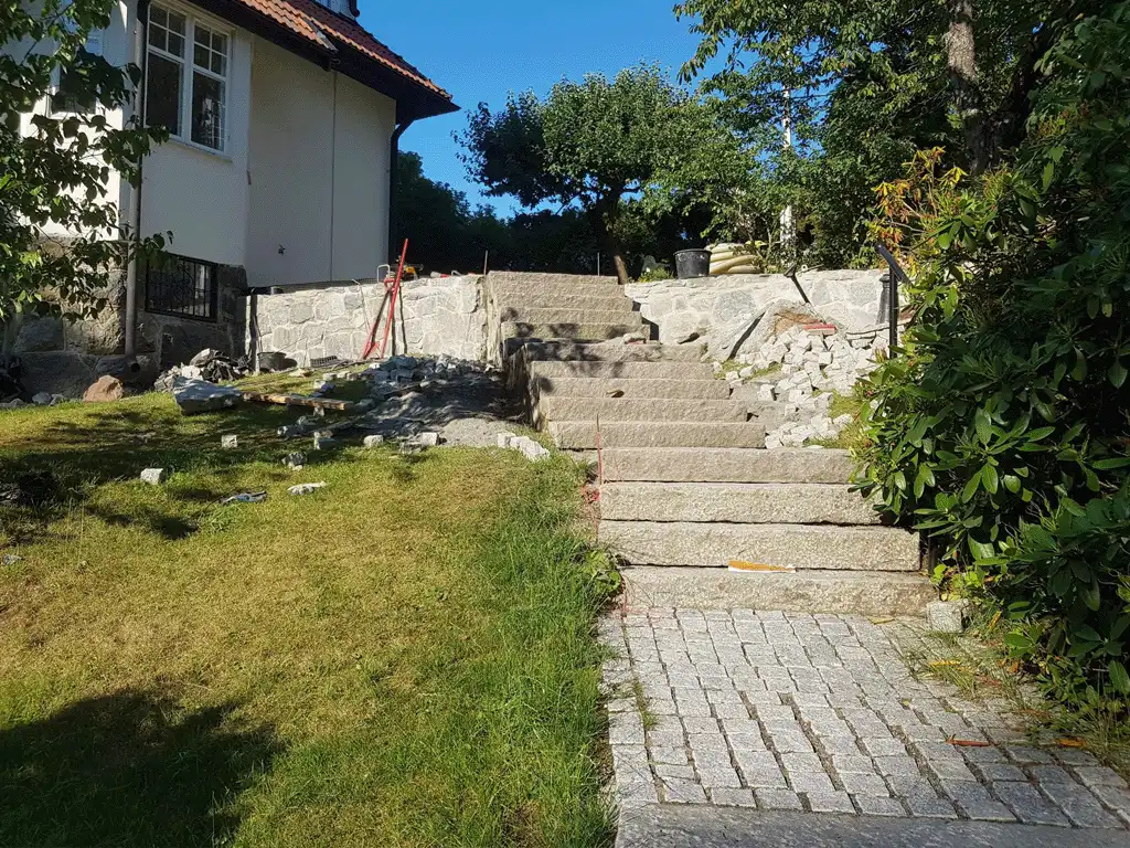 Bilden visar en stentrappa som leder upp till ett hus med en snyggt stenlagd gångväg vid basen. Trappan är byggd av stora, solida stenar och ger både funktionalitet och en rustik charm. Byggmaterial är utspridda runtomkring, vilket visar att projektet fortfarande är under uppbyggnad. Den omgivande gräsmattan är välskött, och träden och grönskan ger en naturlig känsla till scenen. Denna lösning visar ett vackert och hållbart designalternativ för både trappan och gångvägen, som passar väl in i landskapet. Bilden visar en stentrappa som leder upp till ett hus med en snyggt stenlagd gångväg vid basen. Trappan är byggd av stora, solida stenar och ger både funktionalitet och en rustik charm. Byggmaterial är utspridda runtomkring, vilket visar att projektet fortfarande är under uppbyggnad. Den omgivande gräsmattan är välskött, och träden och grönskan ger en naturlig känsla till scenen. Denna lösning visar ett vackert och hållbart designalternativ för både trappan och gångvägen, som passar väl in i landskapet.